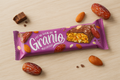 A pink-wrapped Granio date and almond snack bar covered in milk chocolate, surrounded by loose dates and almonds on a wooden background.
