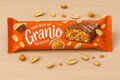 An orange-wrapped Granio peanut butter snack bar covered in milk chocolate, surrounded by peanuts and caramel pieces on a wooden background.