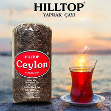 Contextual shot showing the Hilltop Ceylon Black Tea bag next to a steaming glass of red Turkish tea (çay), set against a scenic sunset or seaside background.
