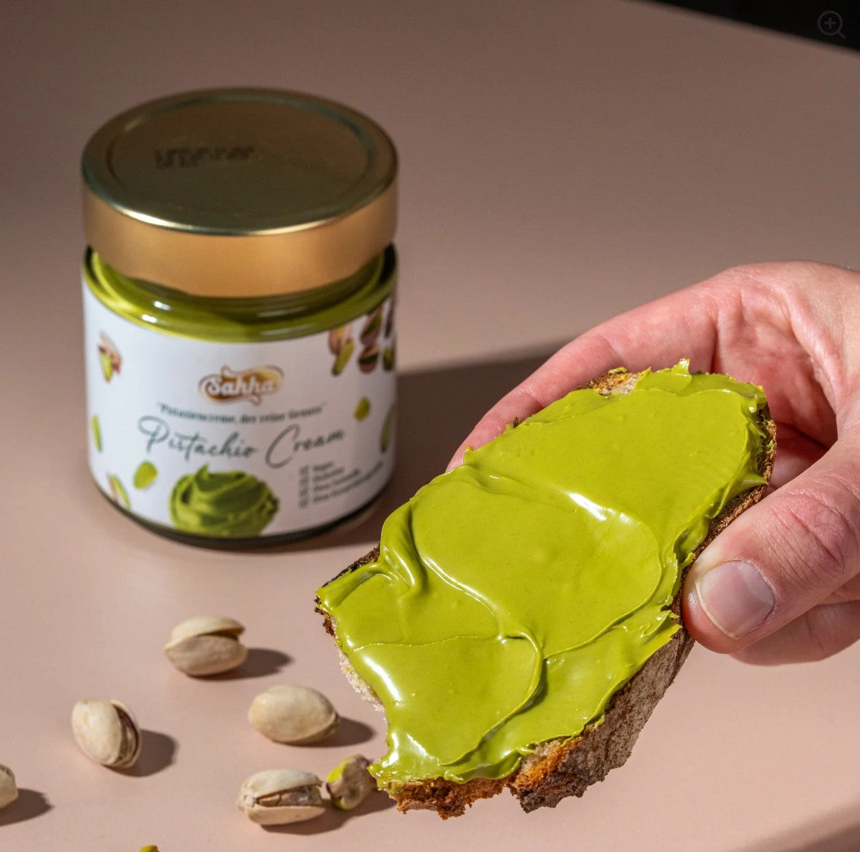 Contextual image of a hand spreading bright green Sahha Pistachio Cream onto a slice of dark bread, with the product jar visible in the background.